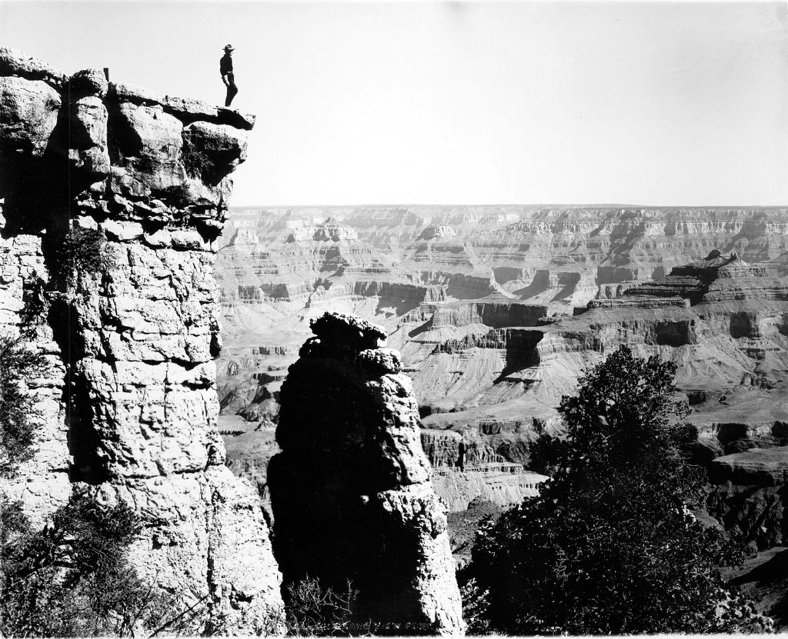 A lone figure stands atop a rocky Grand Canyon overlook in this sweeping vintage black-and-white photograph.