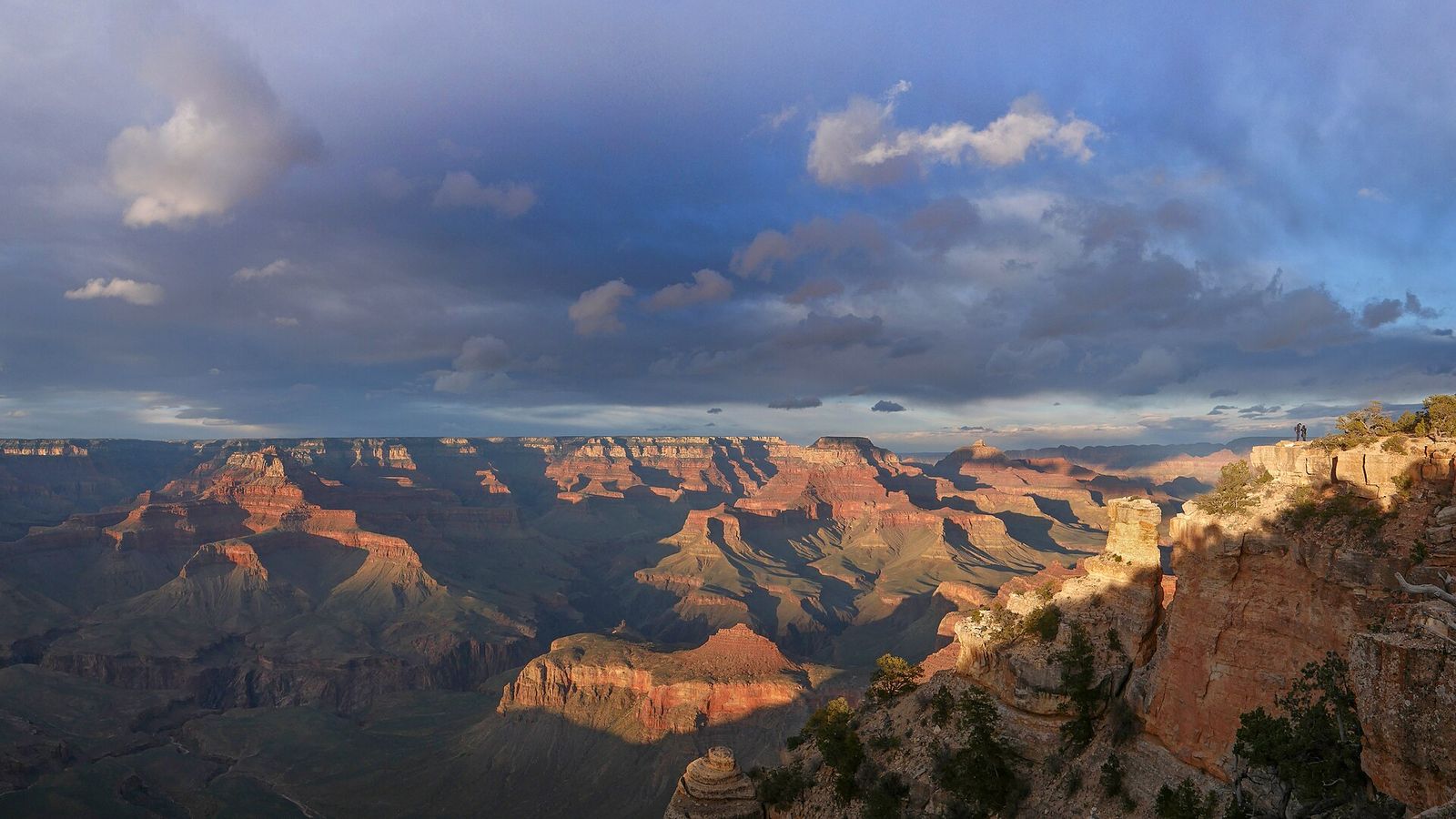Two visitors stand at Yaki Point on the South Rim as dramatic storm clouds sweep over the Grand Canyon's layered buttes.