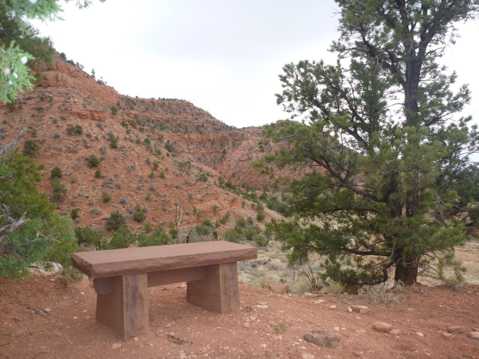 A rustic stone bench sits along a desert hiking trail with red canyon walls and pinyon pines behind it.