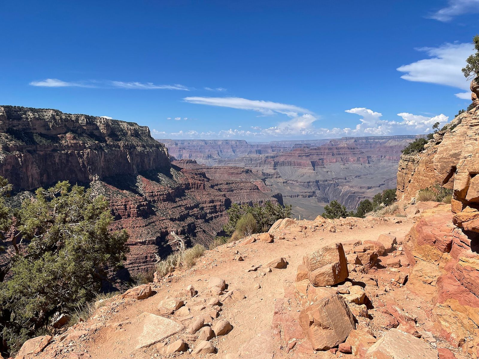 A rocky South Rim overlook reveals sweeping layered canyon walls and deep gorges under a bright blue Arizona sky.