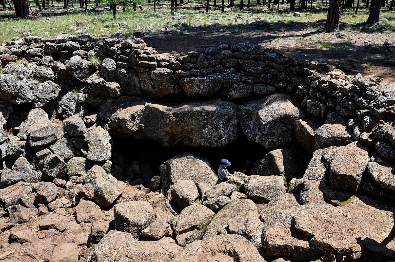 A child in a blue cap explores the rocky entrance of Lava River Cave surrounded by Arizona ponderosa pine forest.