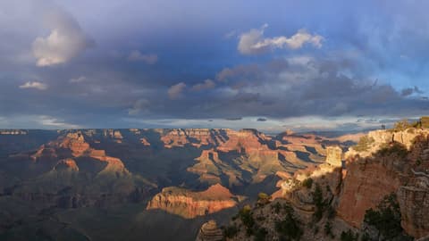 Two visitors stand at Yaki Point on the South Rim as dramatic storm clouds sweep over the Grand Canyon's layered buttes.