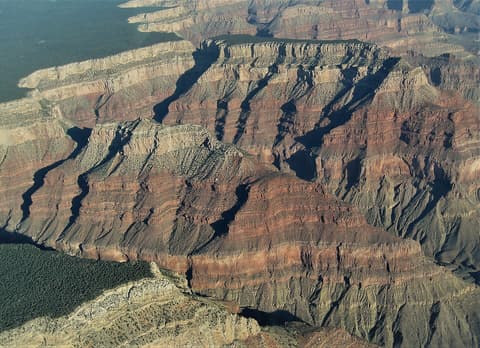 An aerial view of the Grand Canyon's South Rim revealing dramatic layered red rock canyon walls and forested plateaus.