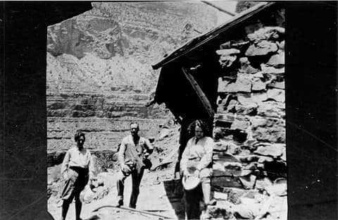 Three early visitors stand beside a rustic stone rest house along the historic Hermit Trail at Grand Canyon.