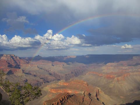 A vivid rainbow arches over the Grand Canyon's layered red rock formations under dramatic storm clouds.