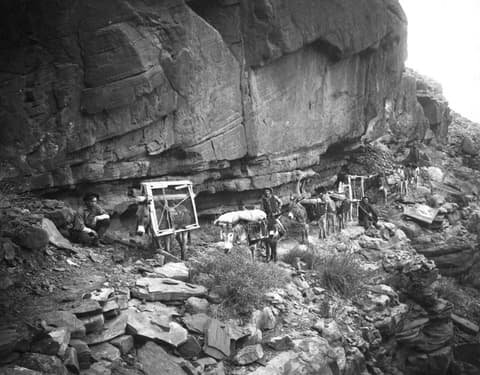 A historic black-and-white photo of pack mules carrying frame loads along a rocky Grand Canyon trail beneath towering canyon walls.