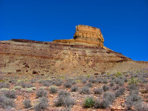 A towering sandstone butte rises above desert scrubland and rocky slopes under a deep blue Arizona sky.