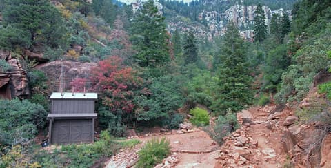 A rustic trail restroom facility sits beside a rocky hiking path surrounded by fall foliage on the North Rim.