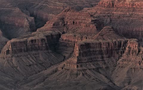 Deeply layered red rock buttes and canyon walls stretch across the Grand Canyon in dramatic shadow and light.
