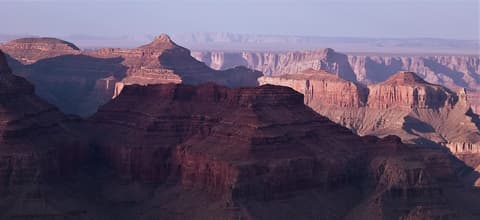 Sweeping Grand Canyon panorama showing layered red rock buttes and deep shadowed gorges under a hazy sky.