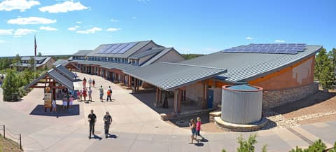 The Grand Canyon South Rim Visitor Center building with solar panels and tourists walking outside on a sunny day.