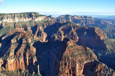 A sweeping aerial view of Grand Canyon's North Rim reveals layered red rock buttes and forested plateaus under clear skies.