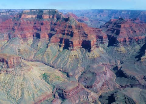 A sweeping aerial panorama of Grand Canyon's layered red rock buttes and deep shadowed canyon walls under blue sky.