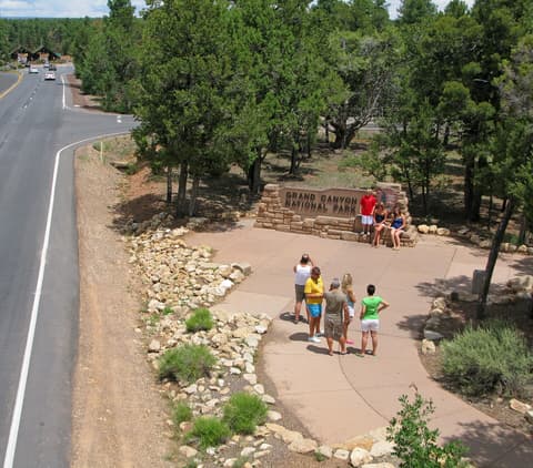 Visitors posing for photos at the Grand Canyon National Park South Rim entrance sign on a sunny day.