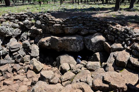 A child in a blue cap explores the rocky entrance of Lava River Cave surrounded by Arizona ponderosa pine forest.