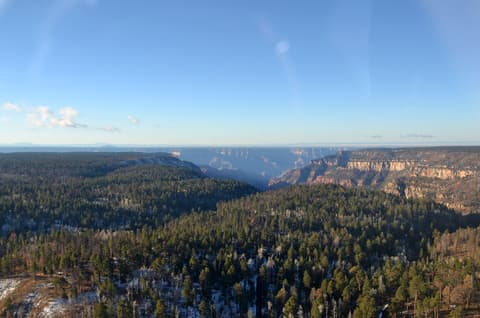 Aerial winter view of the Grand Canyon North Rim with snow-dusted ponderosa pine forest and canyon depths below.